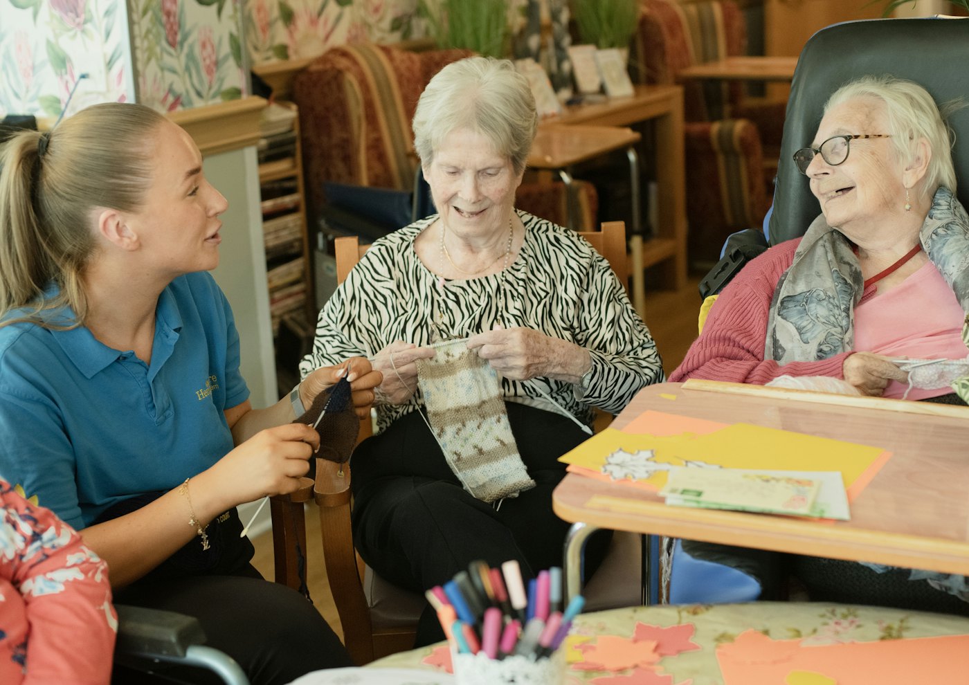 A caregiver helping two older women with a knitting activity
